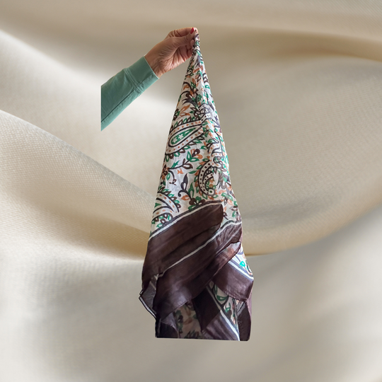 Hand-hemmed lightweight silk scarf with bright paisley pattern held by hand over soft fabric background