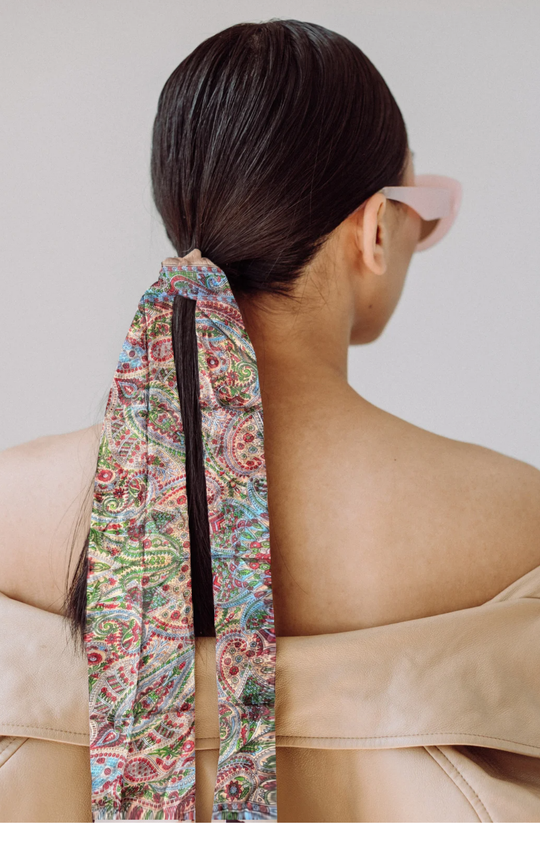 Lightweight hand-hemmed silk scarf in vibrant paisley colors tied in woman's dark hair with beige off-shoulder top and pink sunglasses